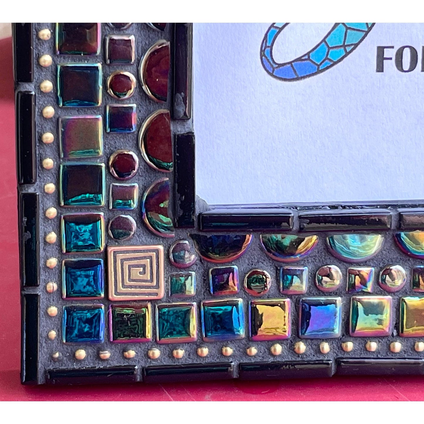 bottom left corner showing close up
5x7 black opal iridescent mosaic tiles with thin black tile outlining the outside as well as the opening for the photo. There are gold square tiles for highlights as well as bronze chain.