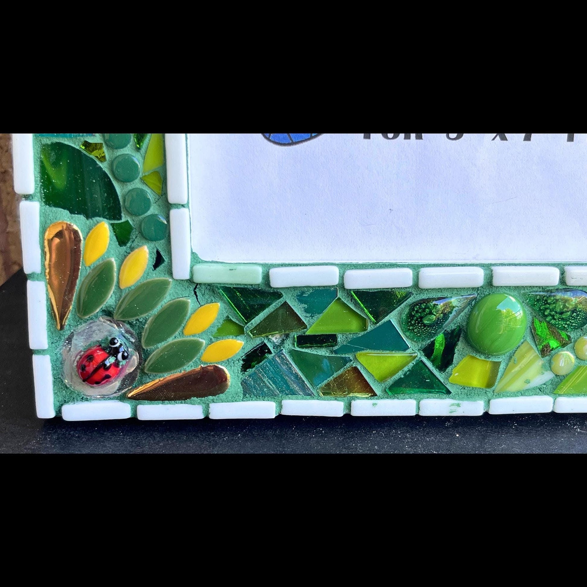 bottom left close up:   5 x 7 Photo frame is shades of green with ceramic leaves, glass beads and tiles. White Thin subway tiles border the inner and outer borders of the frame. Gold Accents and ladybug fused glass in the four corners.  The background grout color is medium green.