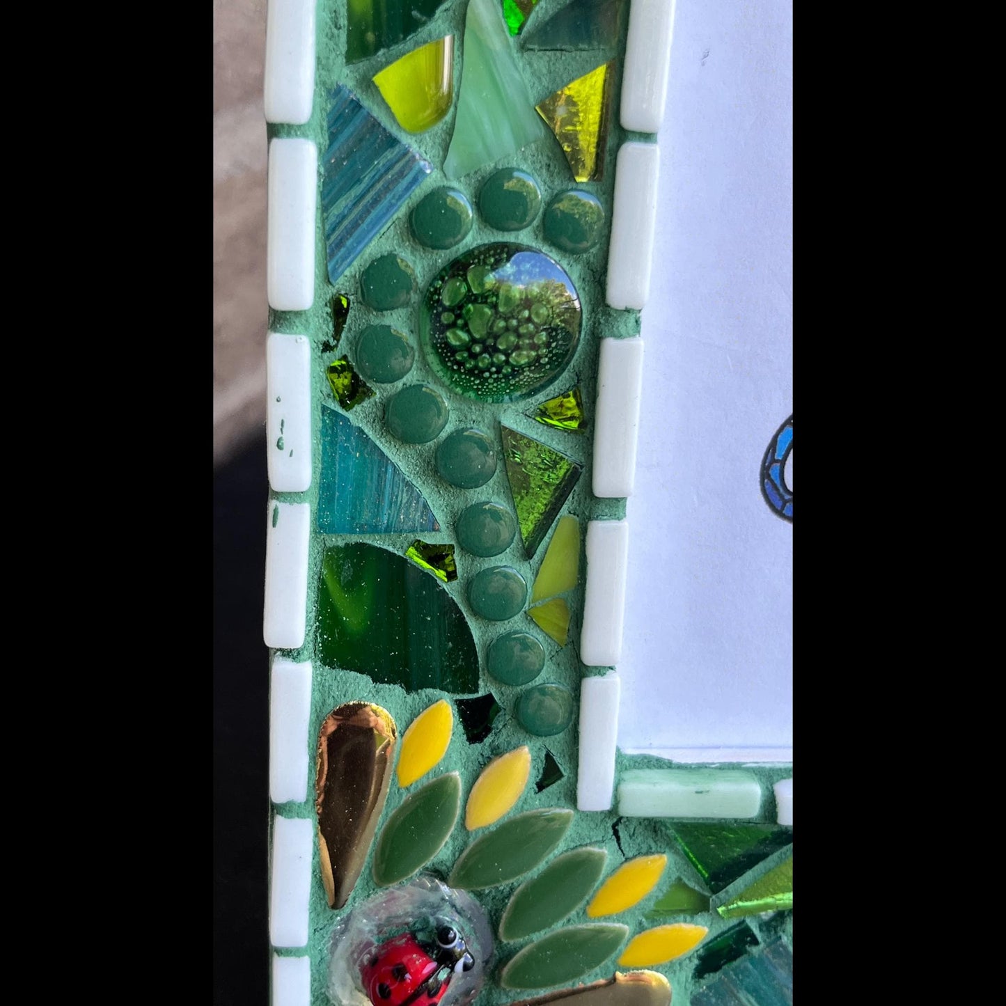 left side close up:
5 x 7 Photo frame is shades of green with ceramic leaves, glass beads and tiles. White Thin subway tiles border the inner and outer borders of the frame. Gold Accents and ladybug fused glass in the four corners.  The background grout color is medium green.
