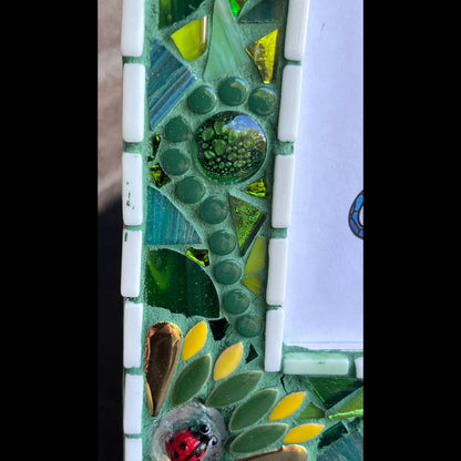 left side close up:
5 x 7 Photo frame is shades of green with ceramic leaves, glass beads and tiles. White Thin subway tiles border the inner and outer borders of the frame. Gold Accents and ladybug fused glass in the four corners.  The background grout color is medium green.