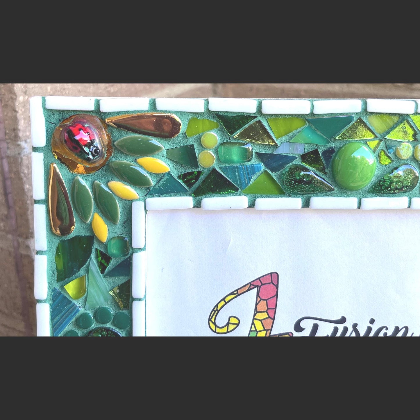 Right top close up:
5 x 7 Photo frame is shades of green with ceramic leaves, glass beads and tiles. White Thin subway tiles border the inner and outer borders of the frame. Gold Accents and ladybug fused glass in the four corners.  The background grout color is medium green.