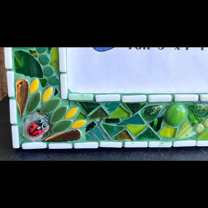 bottom left close up:   5 x 7 Photo frame is shades of green with ceramic leaves, glass beads and tiles. White Thin subway tiles border the inner and outer borders of the frame. Gold Accents and ladybug fused glass in the four corners.  The background grout color is medium green.